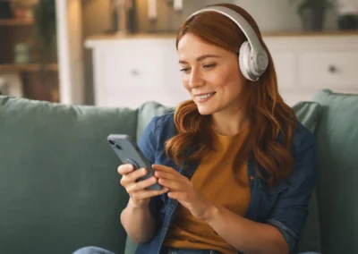En kvinne sitter i sofaen med hodetelefoner på, smiler mens hun ser på mobilen og lytter til musikk eller en podkast i en avslappet hjemmesituasjon.