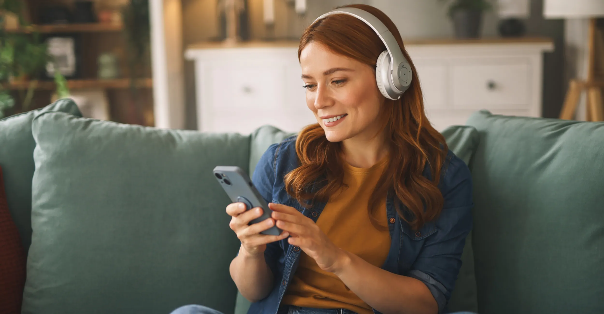 En kvinne sitter i sofaen med hodetelefoner på, smiler mens hun ser på mobilen og lytter til musikk eller en podkast i en avslappet hjemmesituasjon.